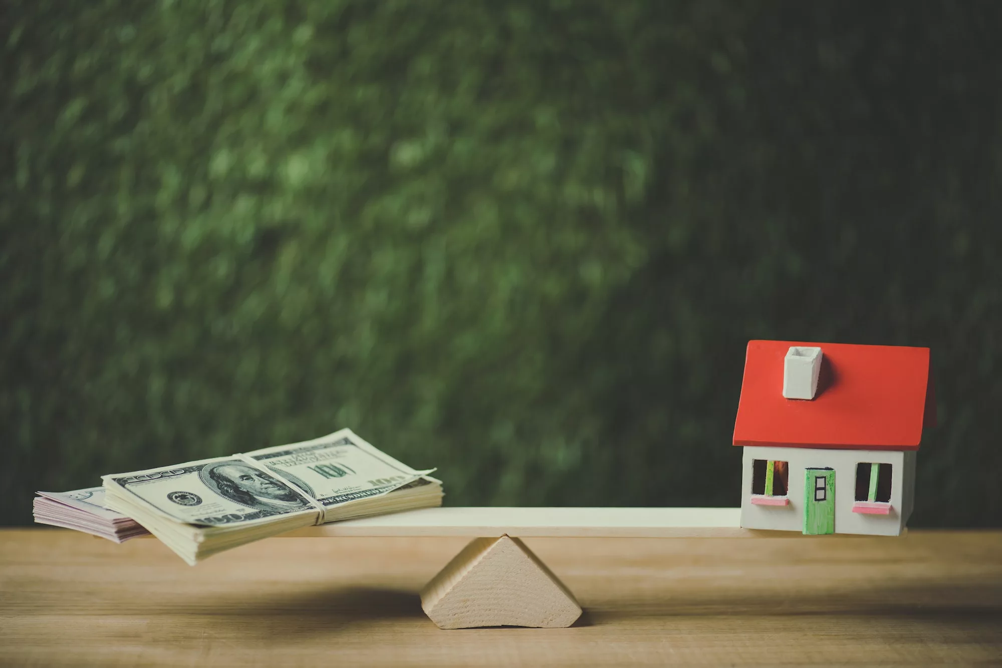dollar banknotes and house model balancing on seesaw on green background